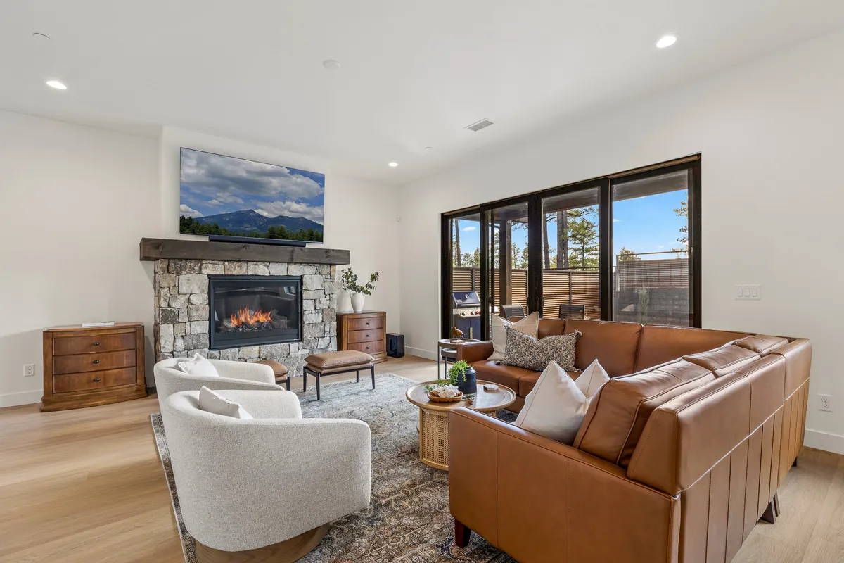 The image focuses on a cozy living room anchored by a fireplace, large windows, a leather sofa, and decorative artwork. Warm tones and soft textures create a welcoming setting for relaxing by the fire or gathering for conversation.