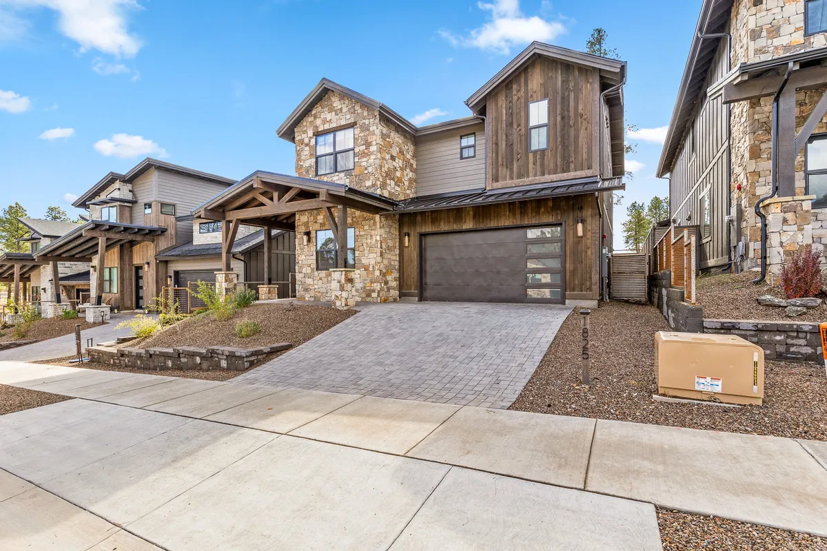 This welcoming exterior image features the home's stone facade, natural wood accents, a spacious driveway, and beautiful landscaping. The inviting porch is perfect for gathering at sunset or preparing for a day of exploration in Flagstaff’s outdoors.