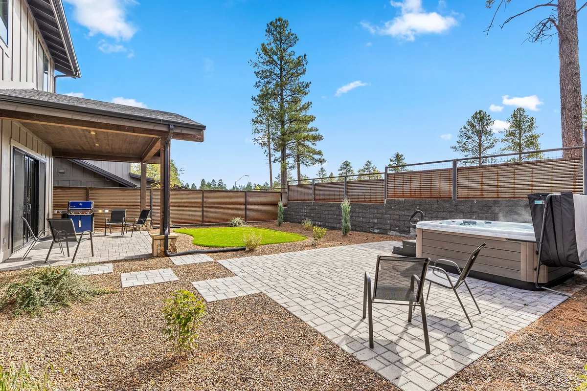 The image showcases the exterior of a modern single-family home surrounded by tall trees, with a spacious patio area, lush lawn, and a steaming hot tub. The sun is setting, casting a warm glow over the scene, ideal for relaxing, socializing, or enjoying the tranquil natural setting.