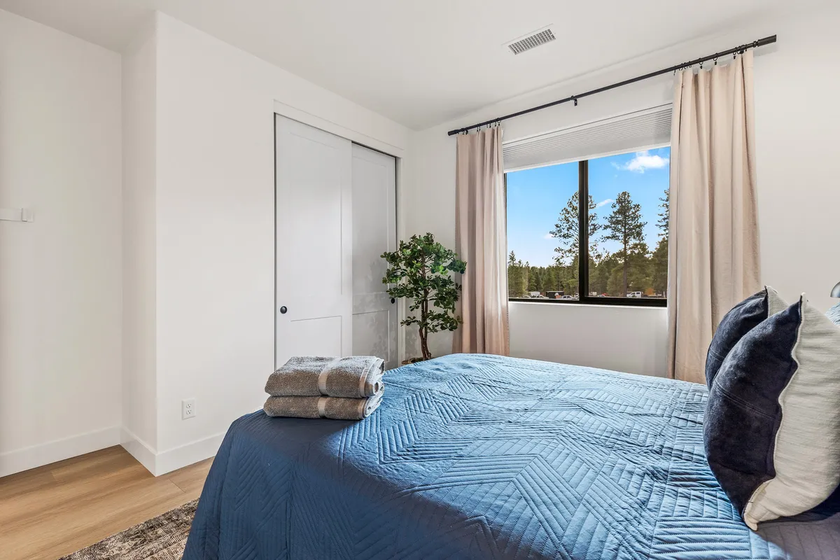 A peaceful bedroom with a bed covered in a blue quilt, an indoor plant, and a window overlooking leafy trees. The window invites natural light and brings attention to the tranquil outdoor setting.