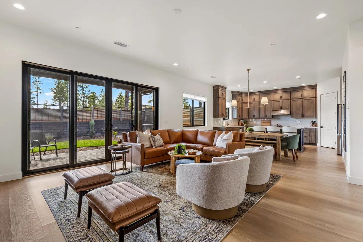 This image captures an inviting open-concept space featuring a leather sofa, round armchairs, a wooden dining table, a visible modern kitchen area, and large windows filling the room with natural light. The arrangement highlights seamless flow between the living, dining, and kitchen zones, with comfortable seating ideal for gatherings.