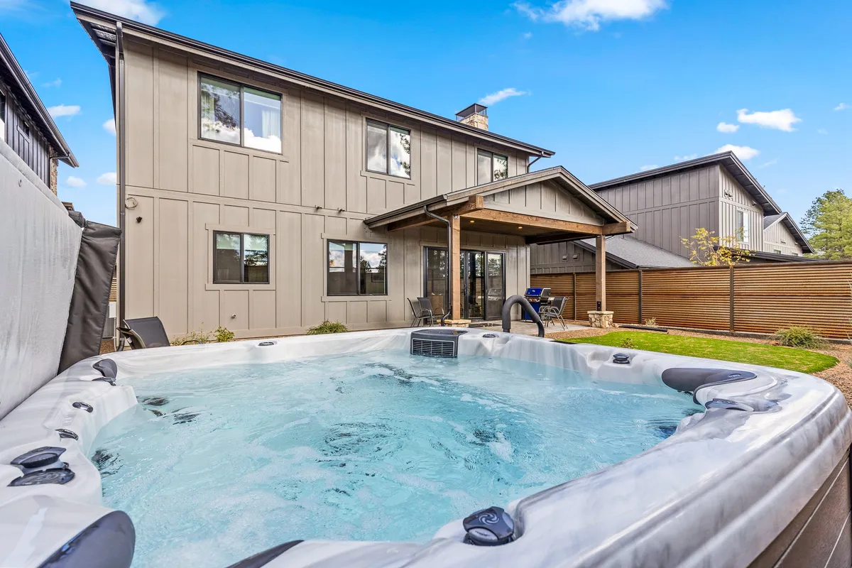 This image features the backyard's inviting hot tub, back patio, the home's exterior facade, and a lush green lawn. The hot tub is the focal point, with comfortable seating nearby and open space perfect for both relaxation and socializing. The evening light hints at sunset, creating a tranquil, magical atmosphere ideal for stargazing or unwinding after a day outdoors.